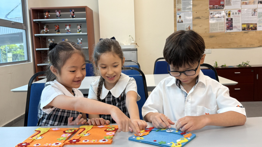 Year 1, primary students sit at a table decorating felt picture frames. Two girls on the left, who have orange frames featuring giraffes, point and look at the blue frame being decorated by the boy on the right. The background features a wooden bulletin board and a shelf displaying Mickey Mouse figures.