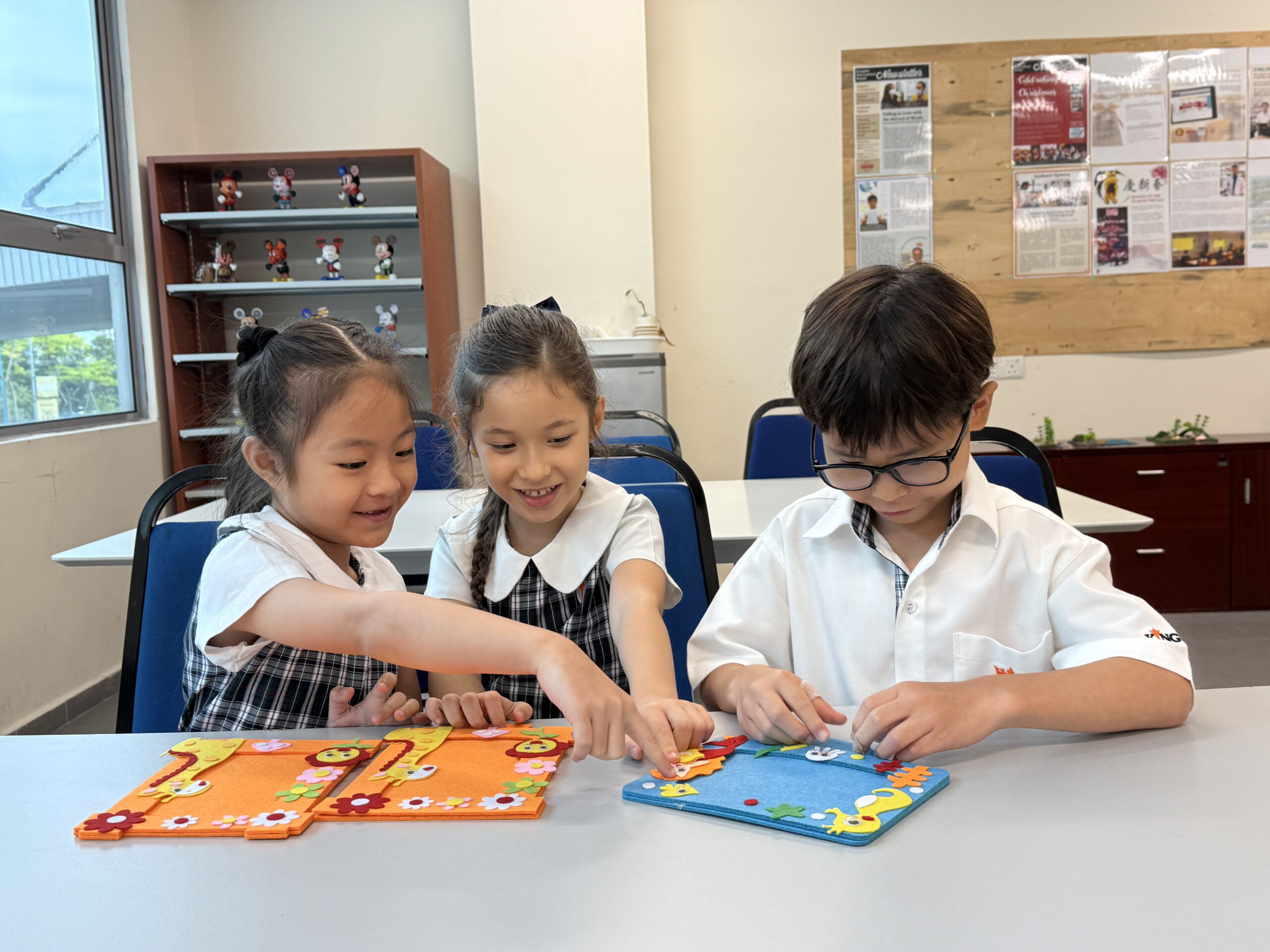Year 1, primary students sit at a table decorating felt picture frames. Two girls on the left, who have orange frames featuring giraffes, point and look at the blue frame being decorated by the boy on the right. The background features a wooden bulletin board and a shelf displaying Mickey Mouse figures.